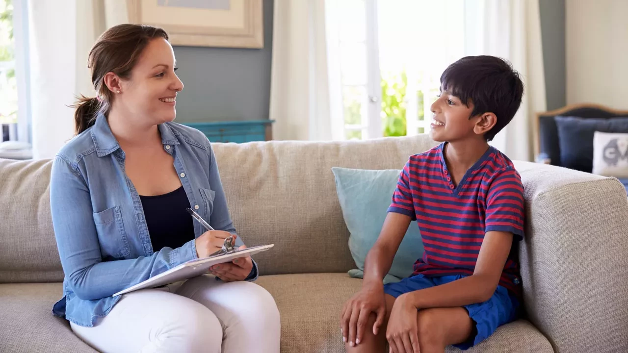 Young boy in a striped shirt sitting on a couch with a therapist in a blue jacket