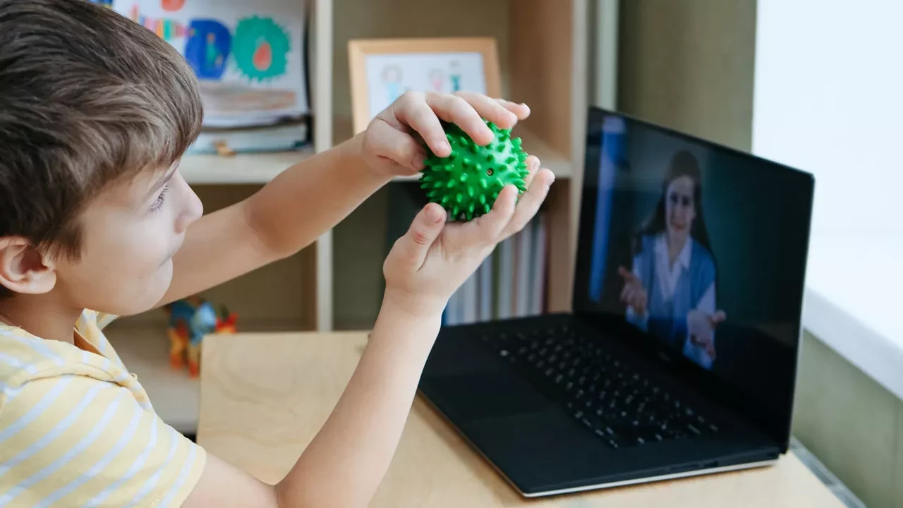 Child playing with a ball in front of a laptop with woman displayed